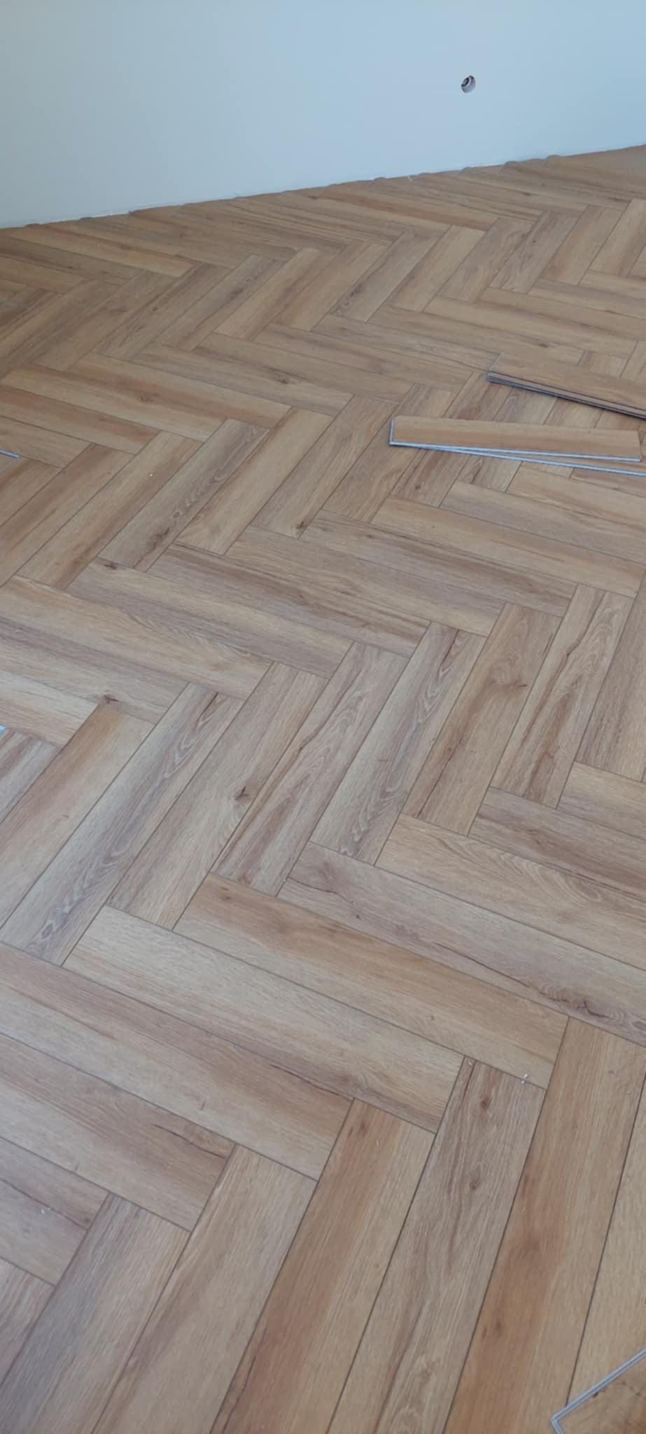 Ukończona podłoga z paneli ułożonych w jodełkę, z widocznymi niedokończonymi panelami i gładką, białą ścianą w tle. Jasne, naturalne oświetlenie.