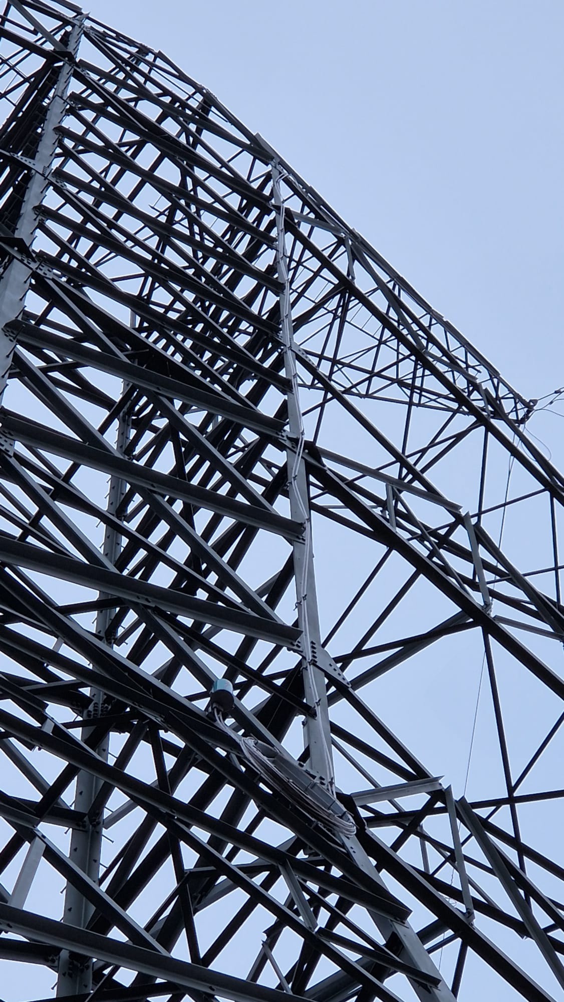 Wysoka, metalowa konstrukcja wieży energetycznej widziana z dołu na tle jasnego nieba. Widoczna skomplikowana siatka belek i wsporników. Prace na wysokości.