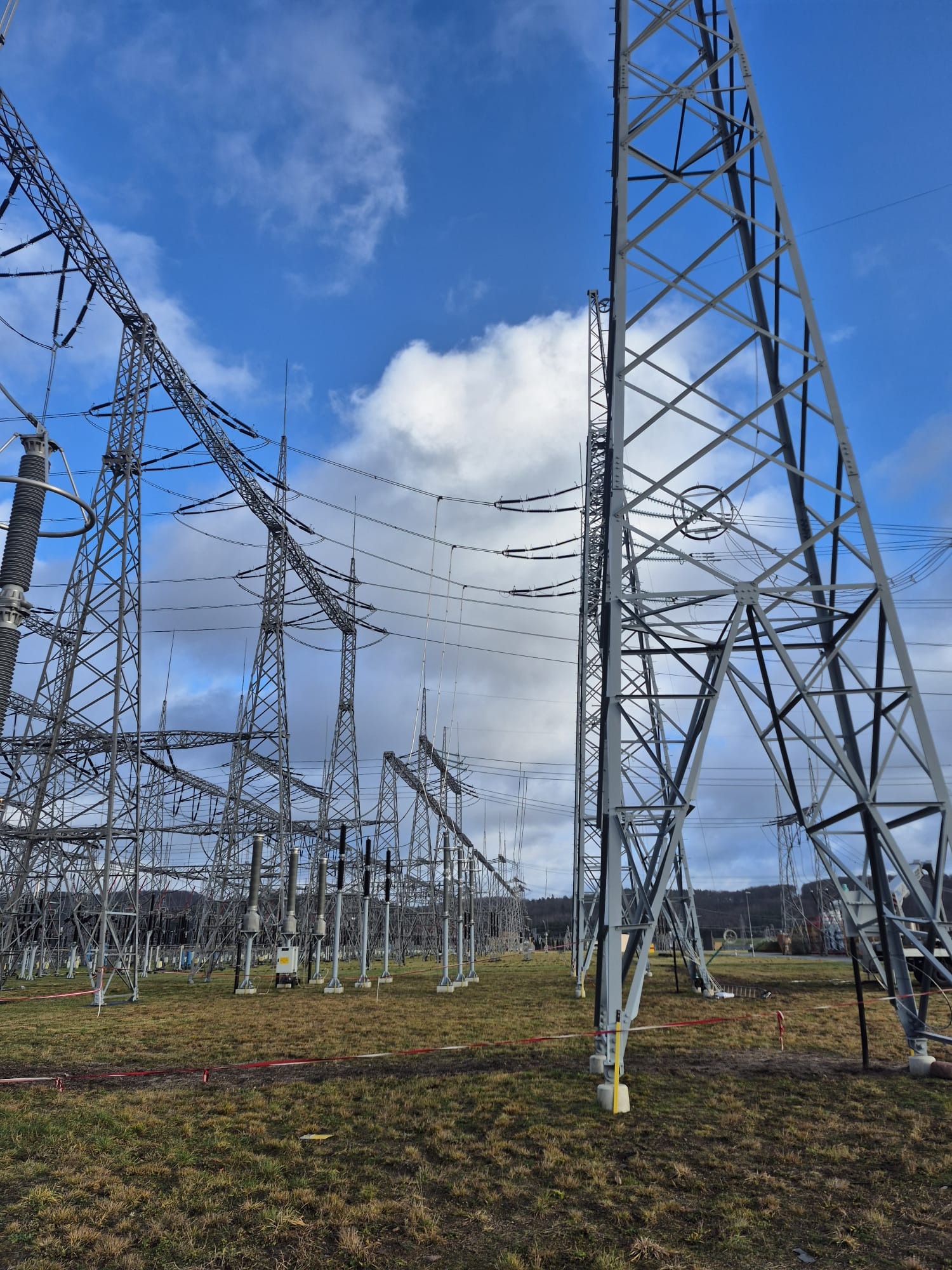 Rozległa stacja transformatorowa z licznymi stalowymi konstrukcjami wspierającymi przewody wysokiego napięcia na tle błękitnego nieba z chmurami. Widok z poziomu gruntu.