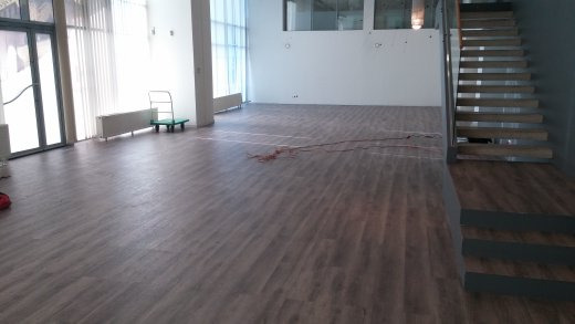 Układanie paneli podłogowych w przestronnym, jasnym wnętrzu z widocznymi przewodami elektrycznymi na podłodze i schodami w tle.