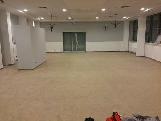 Duża, pusta sala z beżową wykładziną dywanową, białe ściany z szarym pasem, podwójne szklane drzwi w tle, widoczne fragmenty oświetlenia sufitowego i elementy wystroju, w rogu sali biała ścianka...