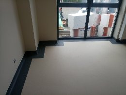 Fragment pomieszczenia z częściowo ułożoną, jasną wykładziną podłogową z ciemnoszarą listwą przypodłogową, widok na okno z materiałami budowlanymi na zewnątrz.