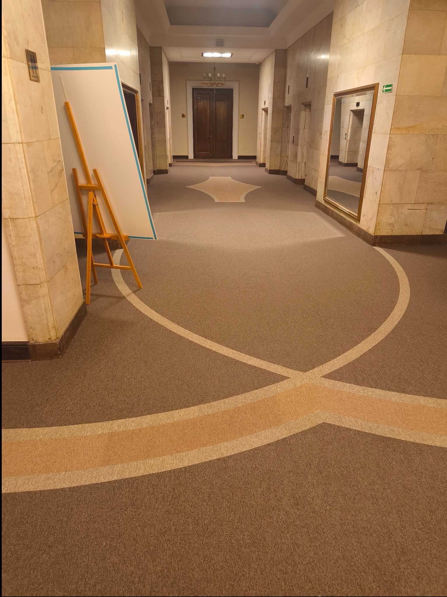 Wzorzysta, brązowa wykładzina w korytarzu z marmurowymi ścianami. Widoczny fragment sztalugi z płótnem i lustro. Perspektywa z dołu podkreśla długość korytarza.