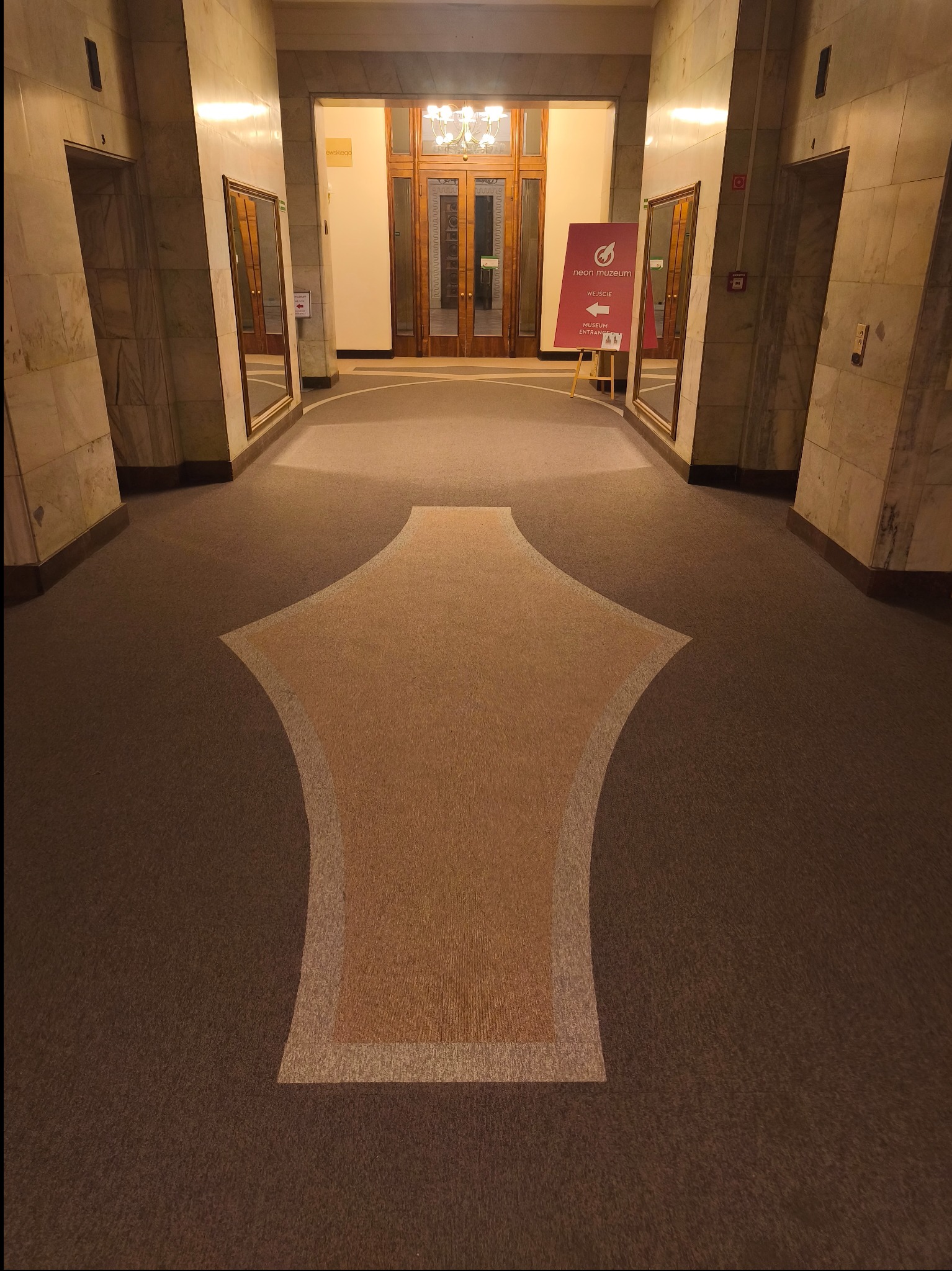 Wzorzysta wykładzina w holu budynku z marmurowymi ścianami i lustrami. Wykładzina z geometrycznym wzorem w odcieniach brązu i beżu prowadzi do dwuskrzydłowych drzwi.