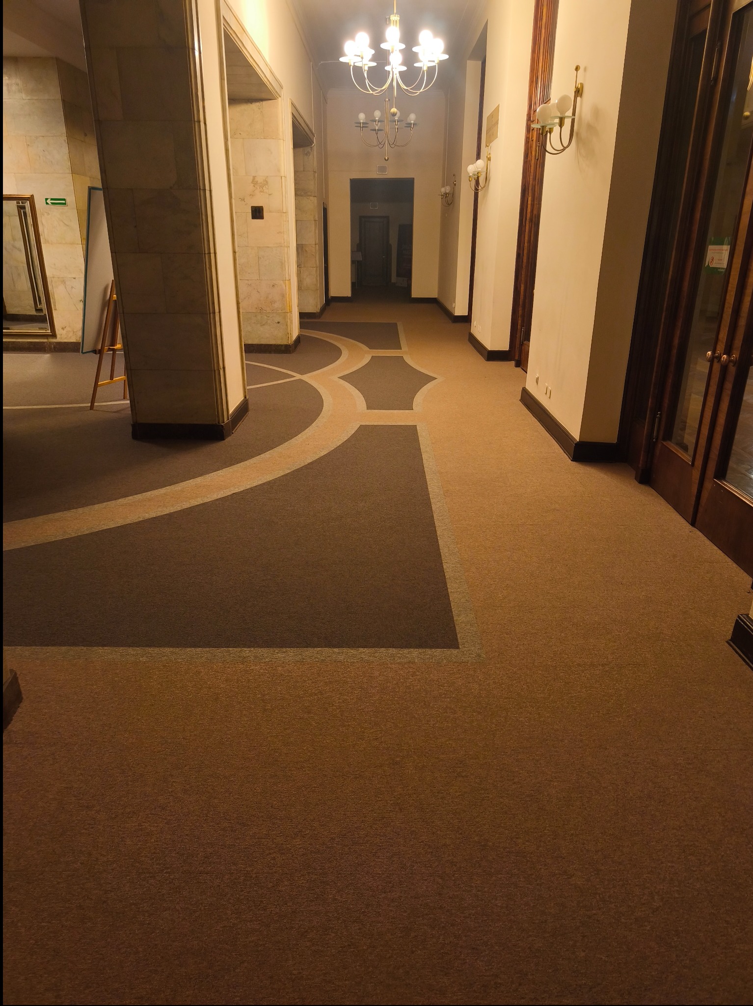 Wzorzysta wykładzina dywanowa w korytarzu z marmurowymi kolumnami i oświetleniem w stylu retro. Brązowa kolorystyka, geometryczne wzory na podłodze.