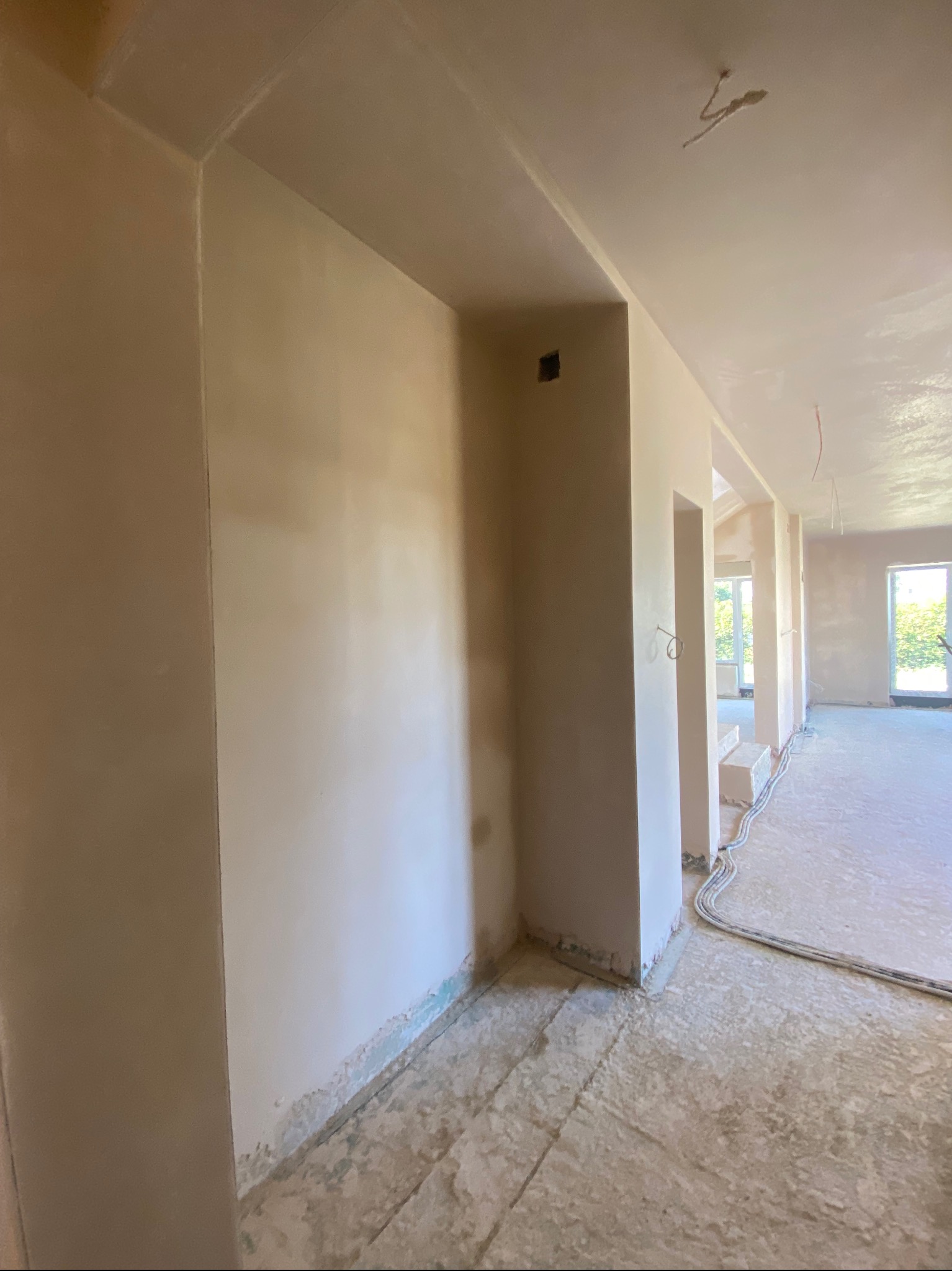 Wnętrze w trakcie prac wykończeniowych: świeżo otynkowane ściany w jasnym odcieniu, widoczne przewody elektryczne przy podłodze, surowy beton na podłodze, ślady po taśmie malarskiej na dole ściany.