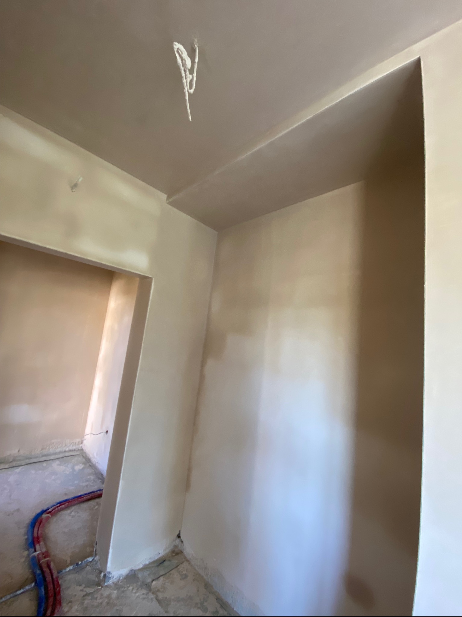 Świeżo otynkowane ściany w pomieszczeniu, widoczne rury w podłodze i sznurek zwisający z sufitu.