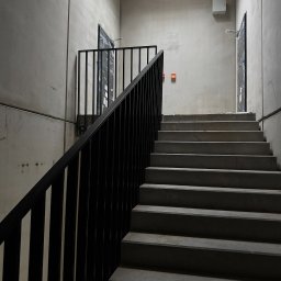 ZAKŁAD PRODUKCJI METALOWEJ BIBI STEEL SPÓŁKA Z OGRANICZONĄ ODPOWIEDZIALNOŚCIĄ - Surowe schody betonowe z czarną, metalową balustradą w minimalistycznym wnętrzu budynku. Widoczne drzwi na każdym poziomie i surowy beton.