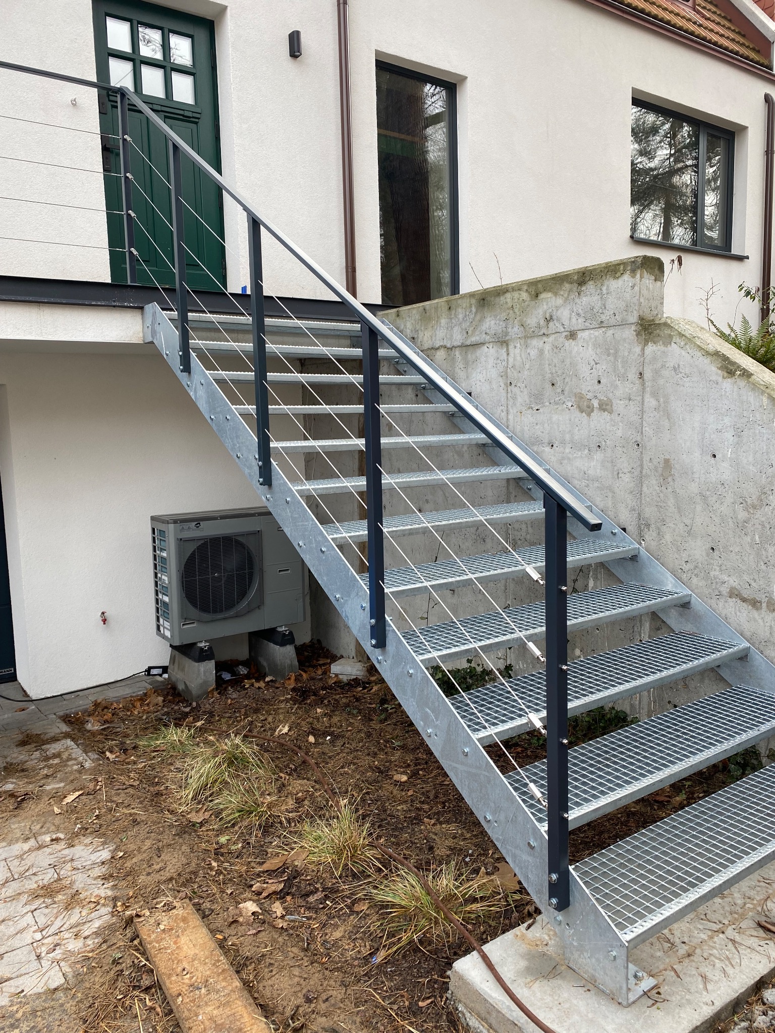 Zewnętrzne schody metalowe z ażurowymi stopniami i czarną balustradą, prowadzące do zielonych drzwi na tle białej ściany budynku z widoczną jednostką zewnętrzną klimatyzacji.