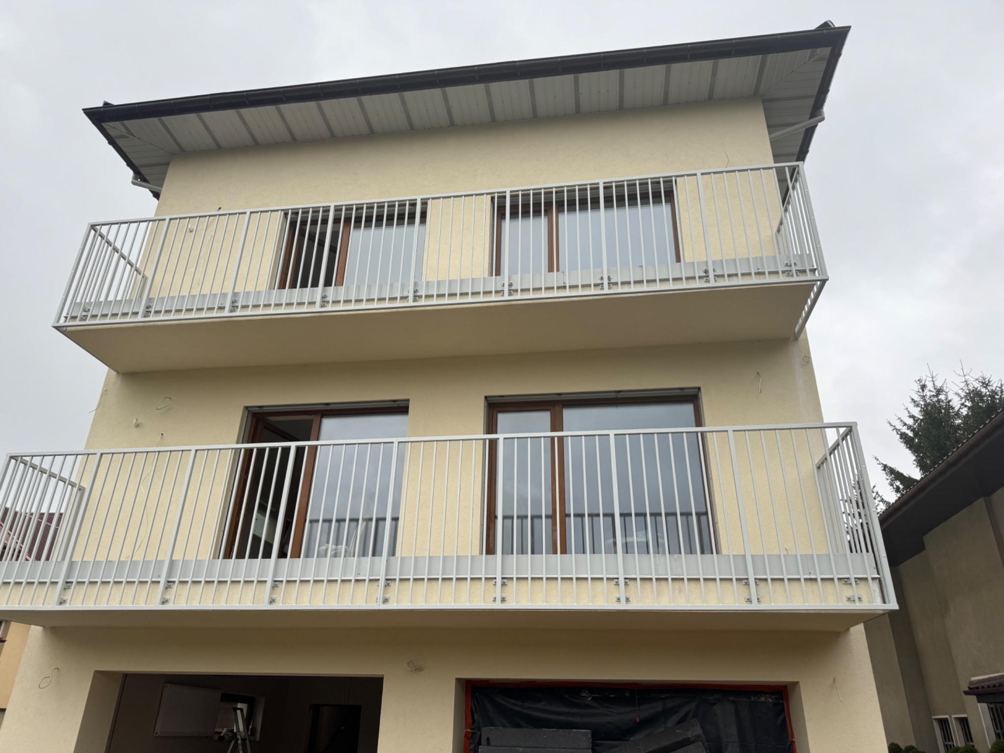 Dwukondygnacyjny budynek w Warszawie z balkonami zabezpieczonymi nowoczesnymi, jasnymi balustradami z pionowych prętów. Widoczne brązowe ramy okienne i szary, pochmurny dzień.