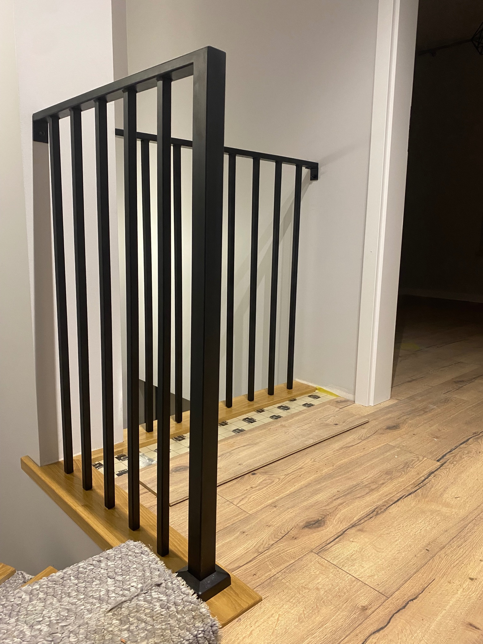 Częściowo zamontowana czarna balustrada z pionowymi prętami na drewnianych schodach z dywanikiem, widoczne podkłady pod panele podłogowe.