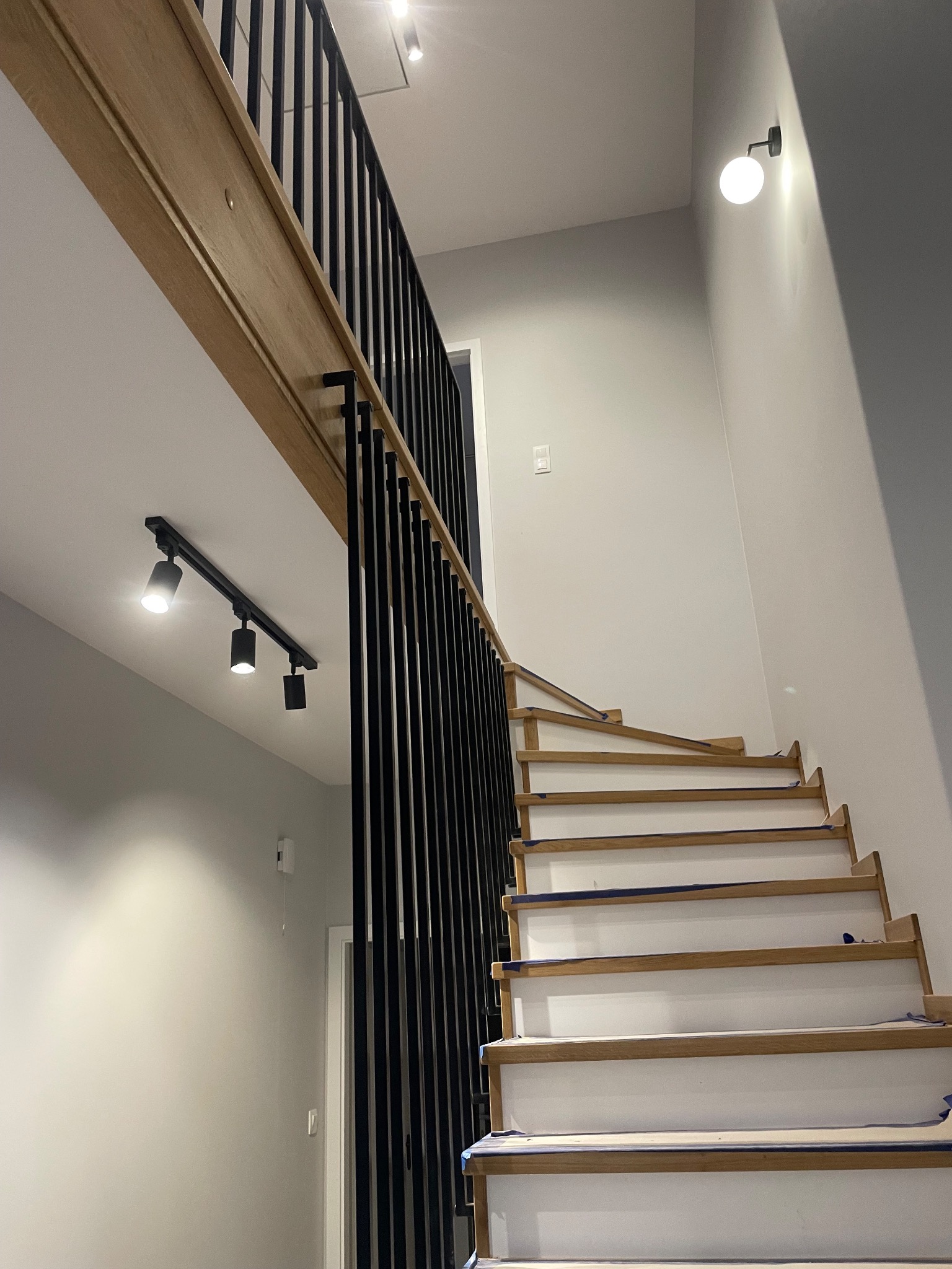 Wnętrze z drewnianymi schodami w trakcie wykończenia, zabezpieczone taśmą malarską, z czarną metalową balustradą i oświetleniem punktowym.