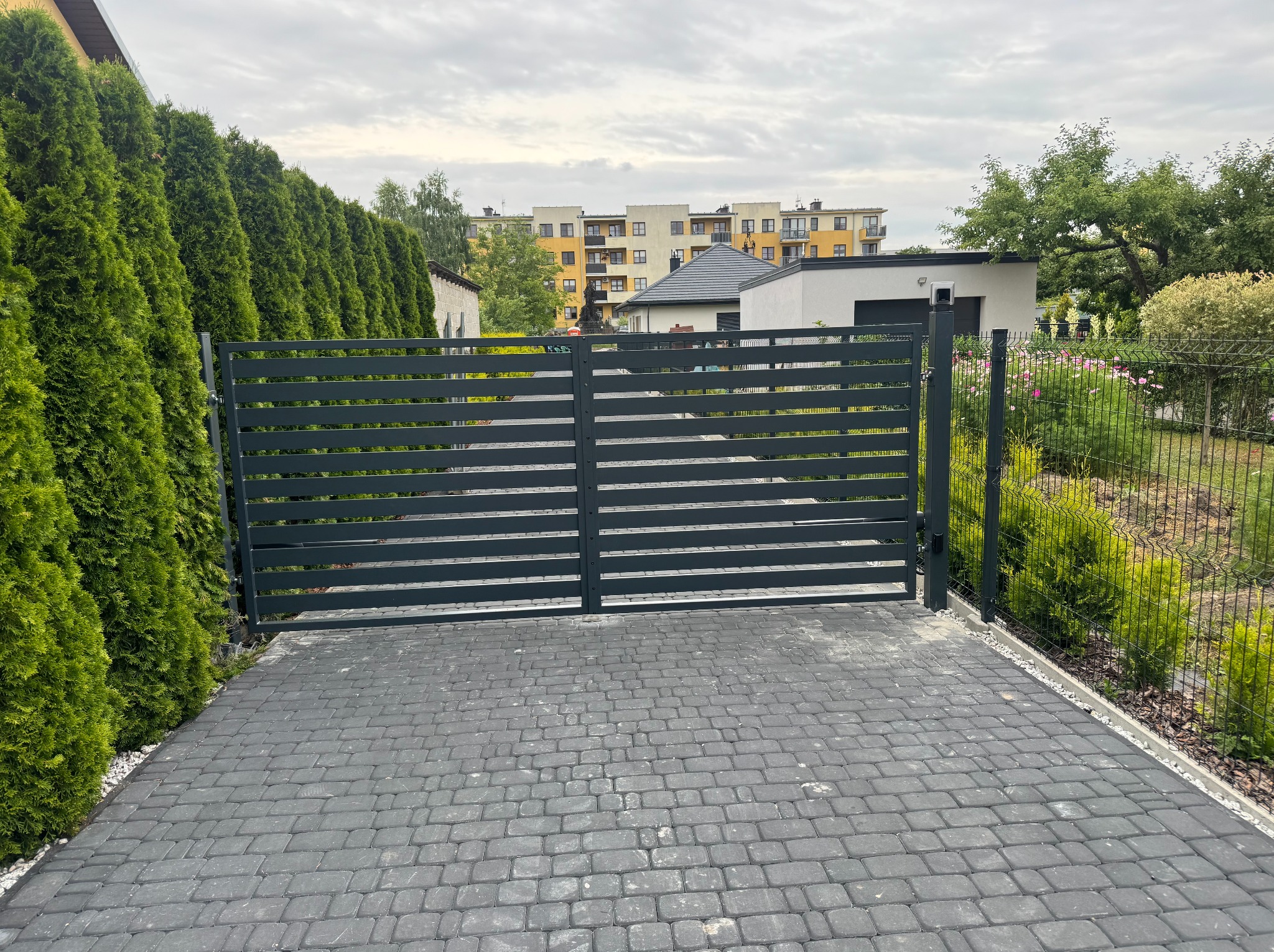 Dwuskrzydłowa brama wjazdowa z poziomymi profilami, pomalowana na szaro, na tle brukowanej nawierzchni i zielonych tuj. W tle budynki mieszkalne i domy.