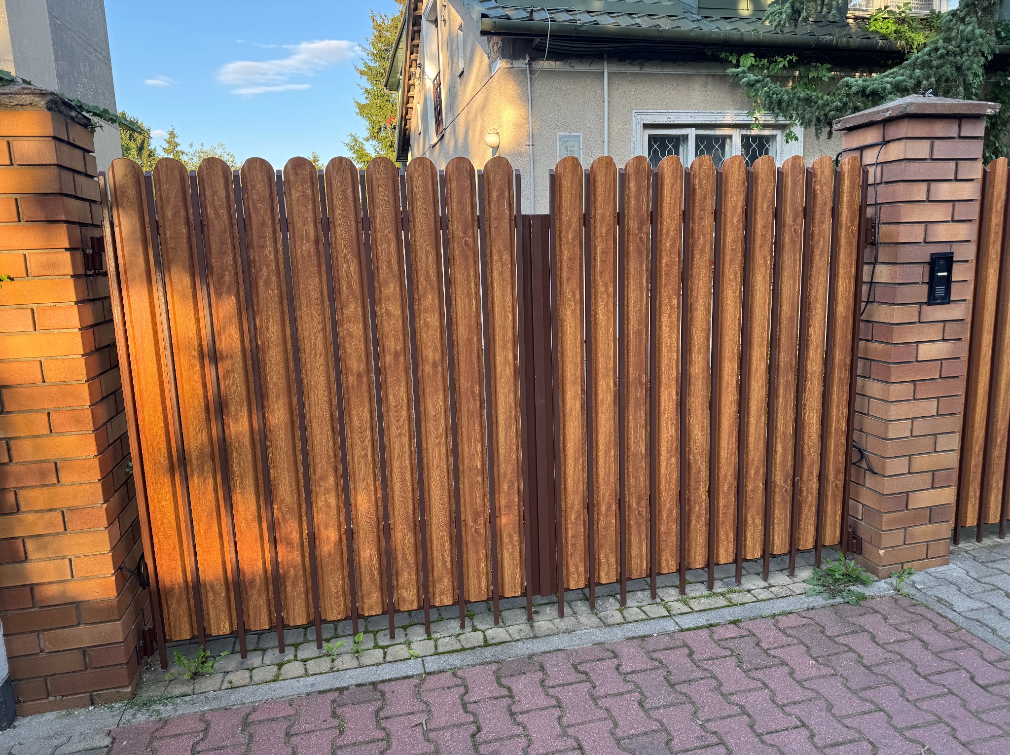 Aluminiowe panele imitujące drewno w ogrodzeniu posesji w Warszawie. Elementy pionowe z zaokrąglonym wykończeniem, zamontowane na metalowej konstrukcji.