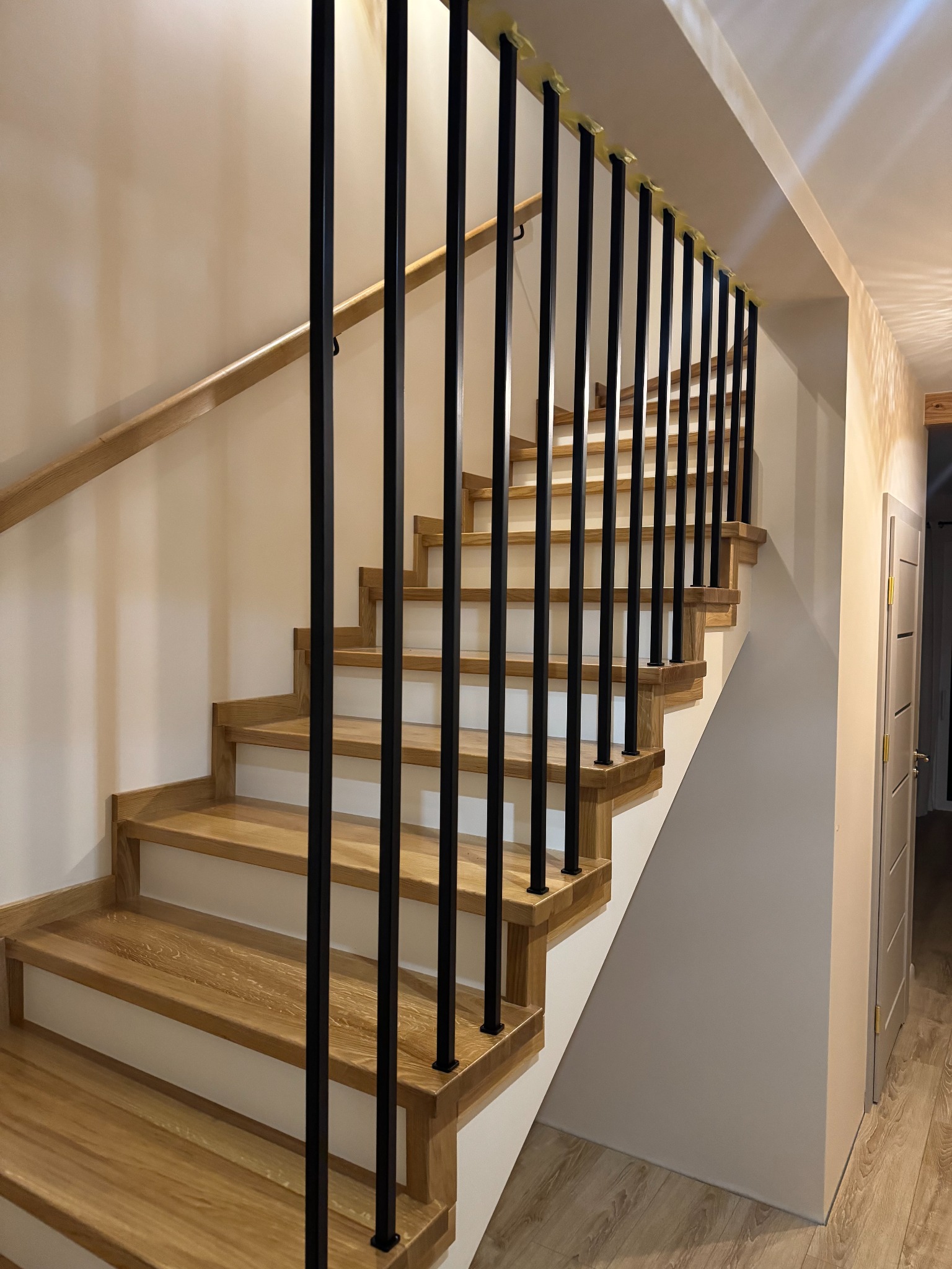 Drewniane schody z czarnymi, metalowymi słupkami i jasną, drewnianą poręczą. Widok z dołu, oświetlenie naturalne, minimalistyczny design.