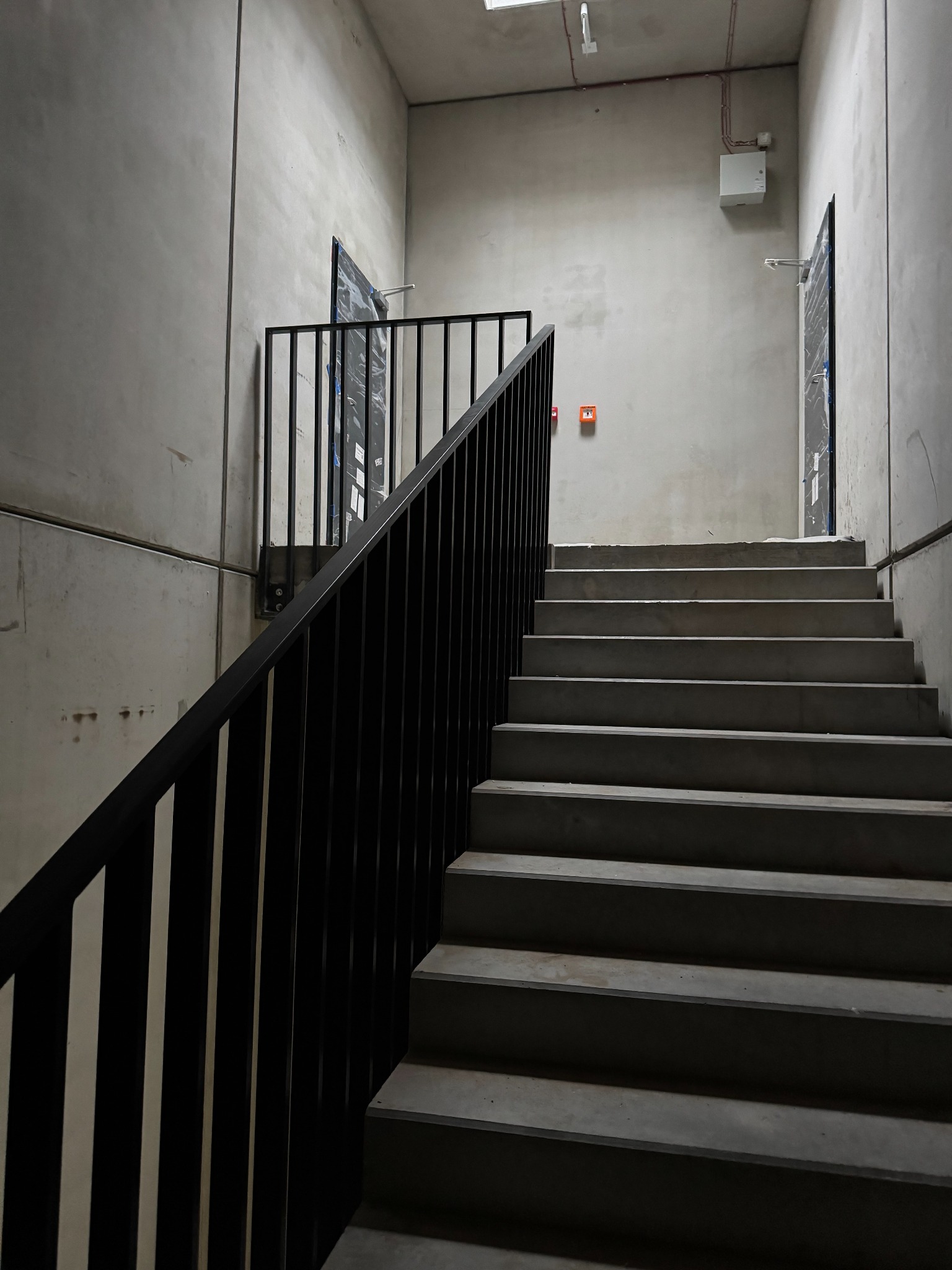 Surowe schody betonowe z czarną, metalową balustradą w minimalistycznym wnętrzu budynku. Widoczne drzwi na każdym poziomie i surowy beton.