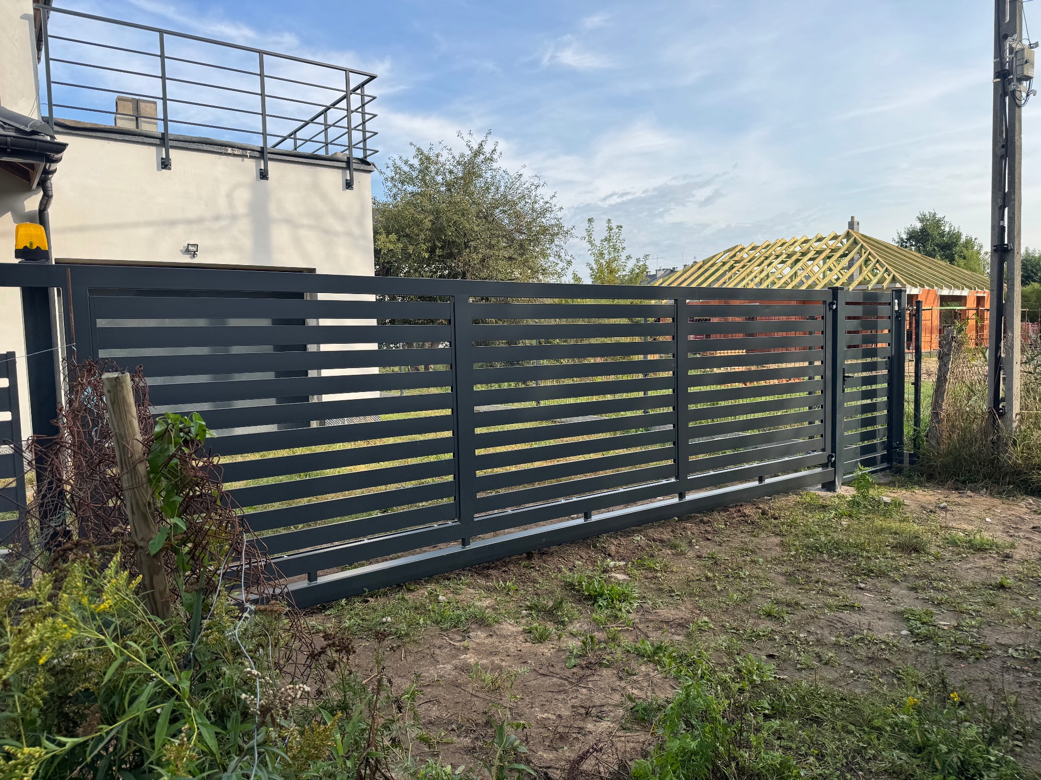 Antracytowa brama przesuwna ze stali węglowej, ocynkowana ogniowo, z furtką i balustradą balkonową. Całość lakierowana proszkowo, na tle budowy domu.