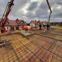 MARKON ADRIAN KONIECZNY - Zbrojenie płyty fundamentowej przygotowane do zalania betonem, widoczne pompy do betonu, mieszalnik i ekipa budowlana na tle domów jednorodzinnych.