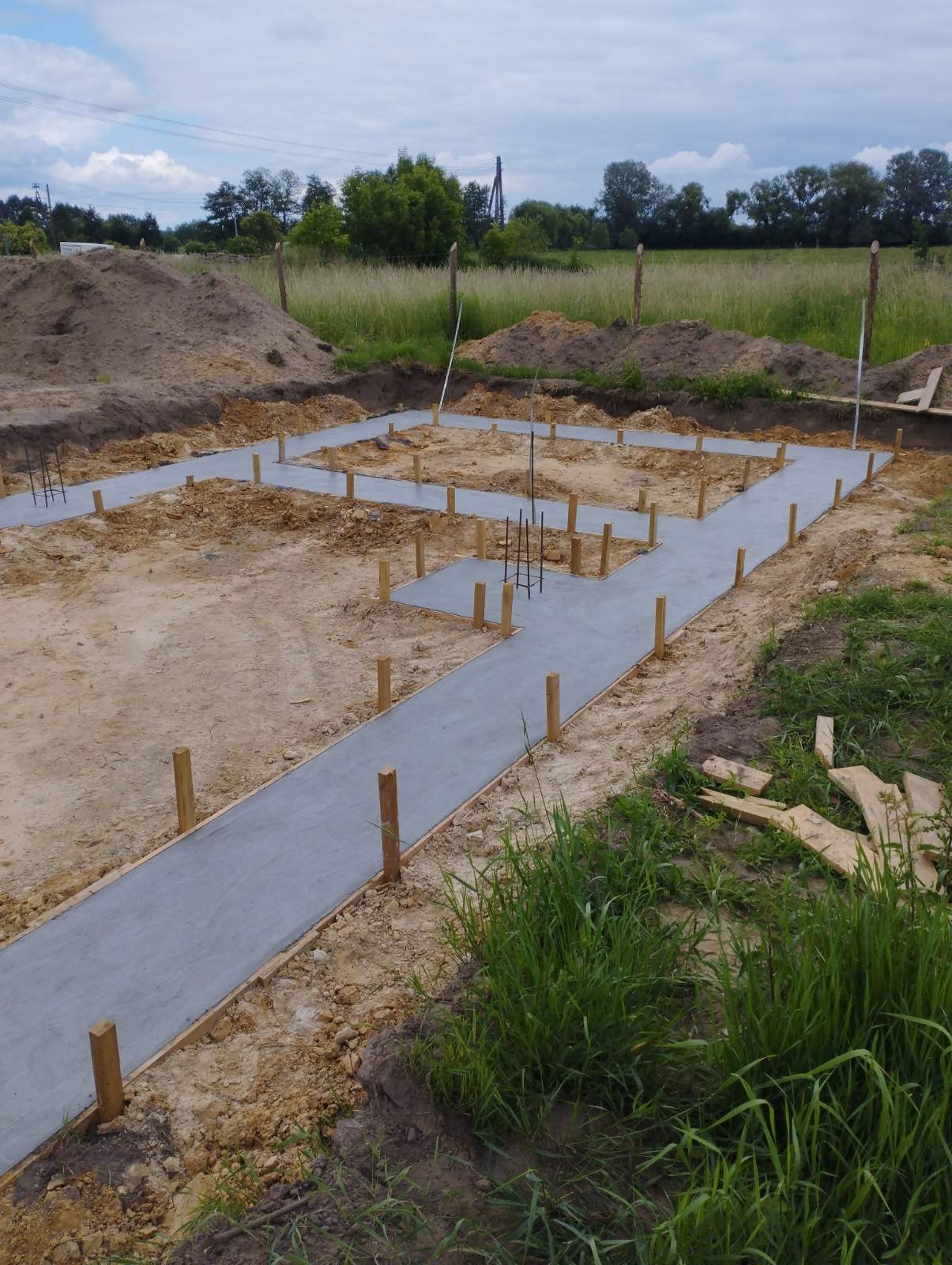 Zalane betonem, drewniane szalunki fundamentów domu jednorodzinnego na działce z ziemią i trawą, widoczne pionowe pręty zbrojeniowe.