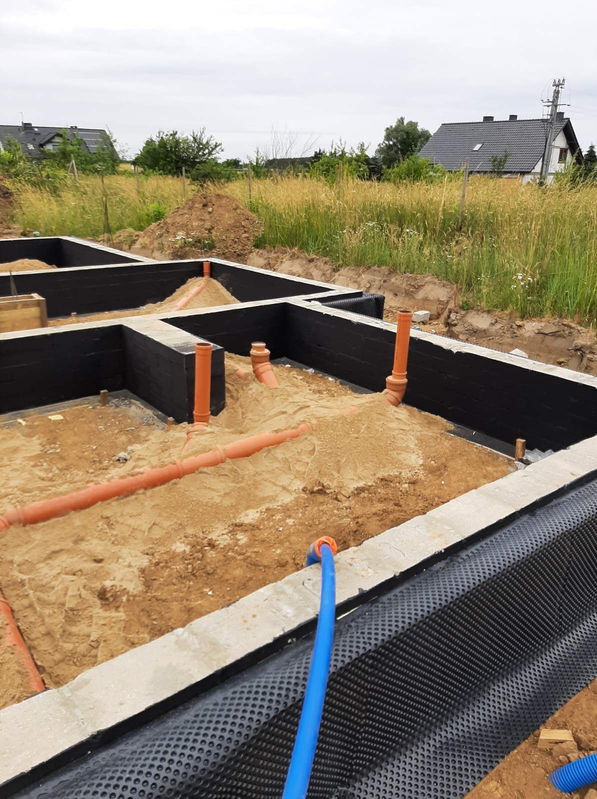 Wykop pod fundamenty z izolacją przeciwwilgociową, wystającymi rurami kanalizacyjnymi i zasypanym piaskiem, widok na budynki mieszkalne w tle.