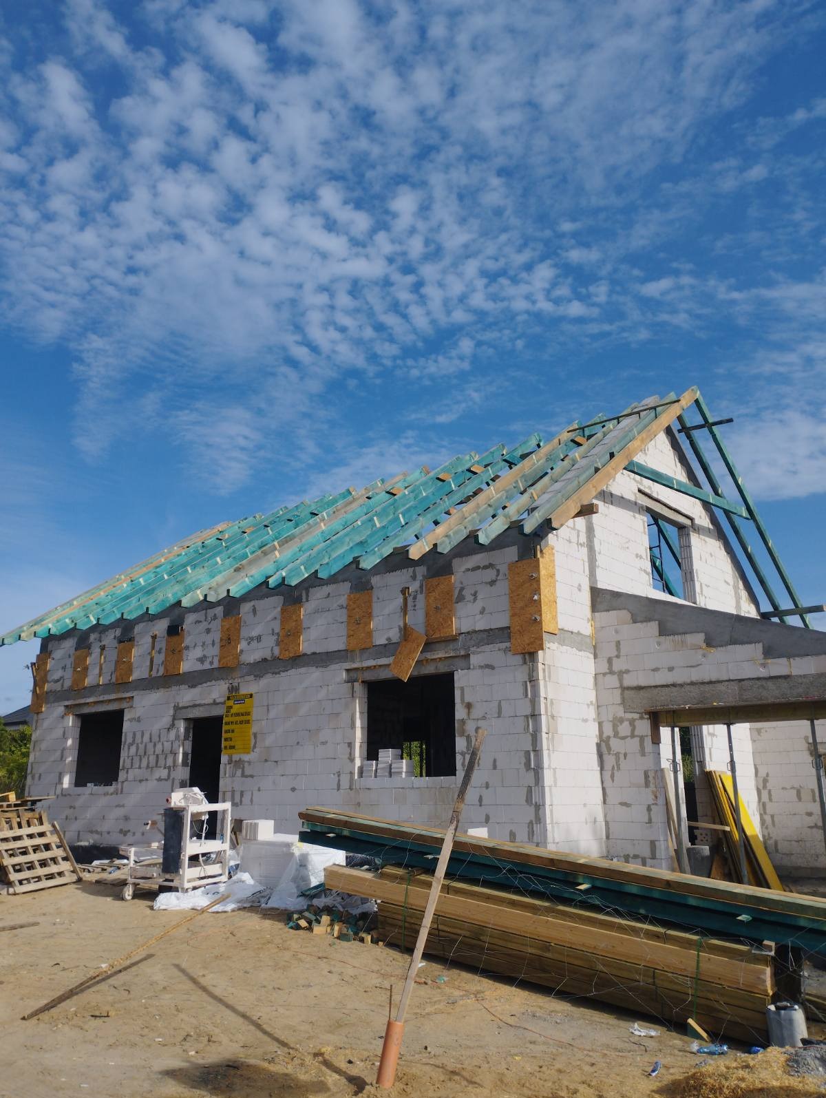 Surowy budynek z białego bloczka w trakcie budowy, z widoczną konstrukcją dachu pokrytą zieloną folią, oraz przygotowanymi deskami na pierwszym planie i błękitnym niebem w tle.