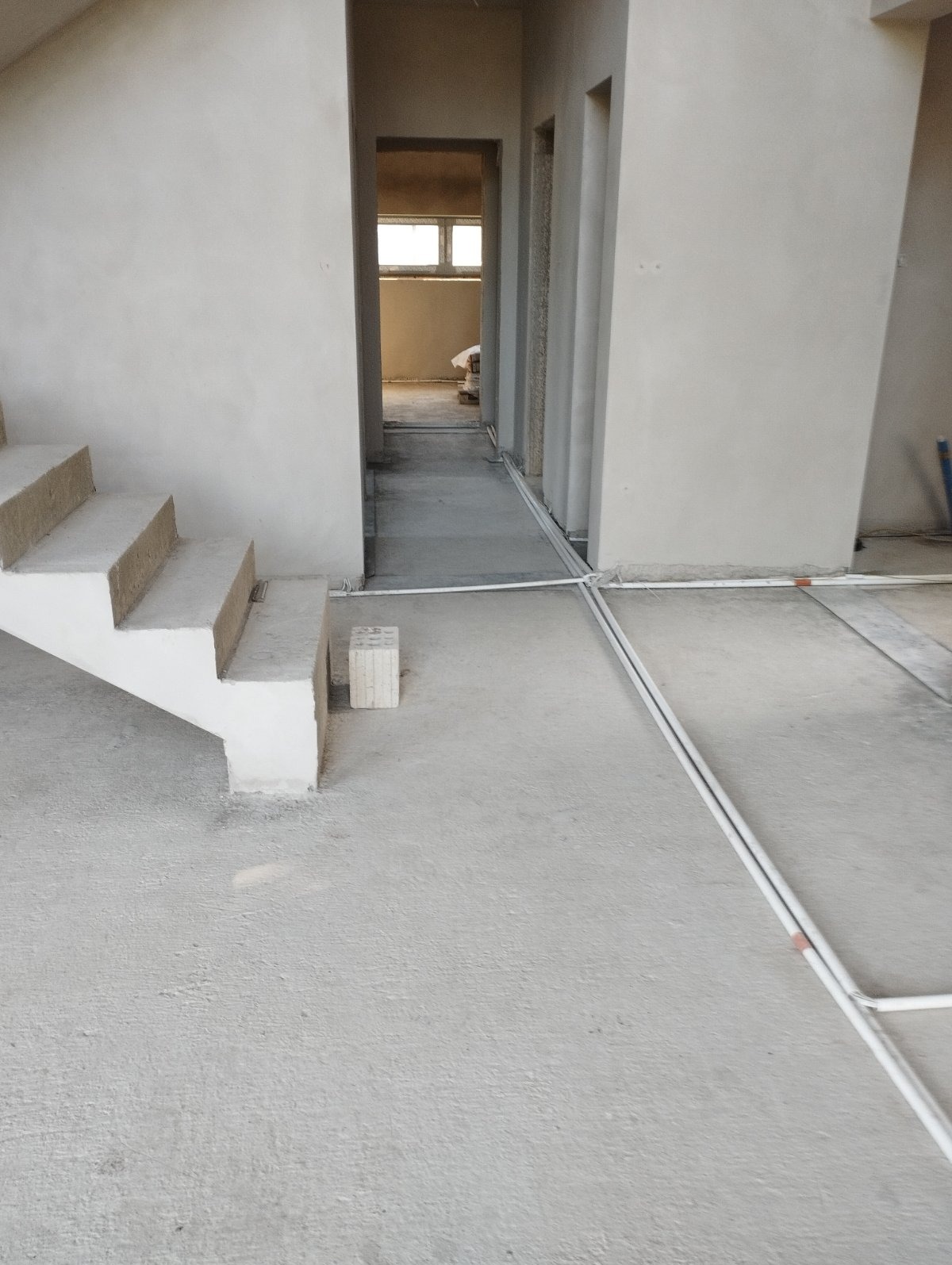 Surowy stan wnętrza domu w trakcie budowy: betonowa podłoga, schody w stanie deweloperskim, ściany pokryte szarym tynkiem, widoczne rury instalacji na podłodze, w głębi widok na okno.