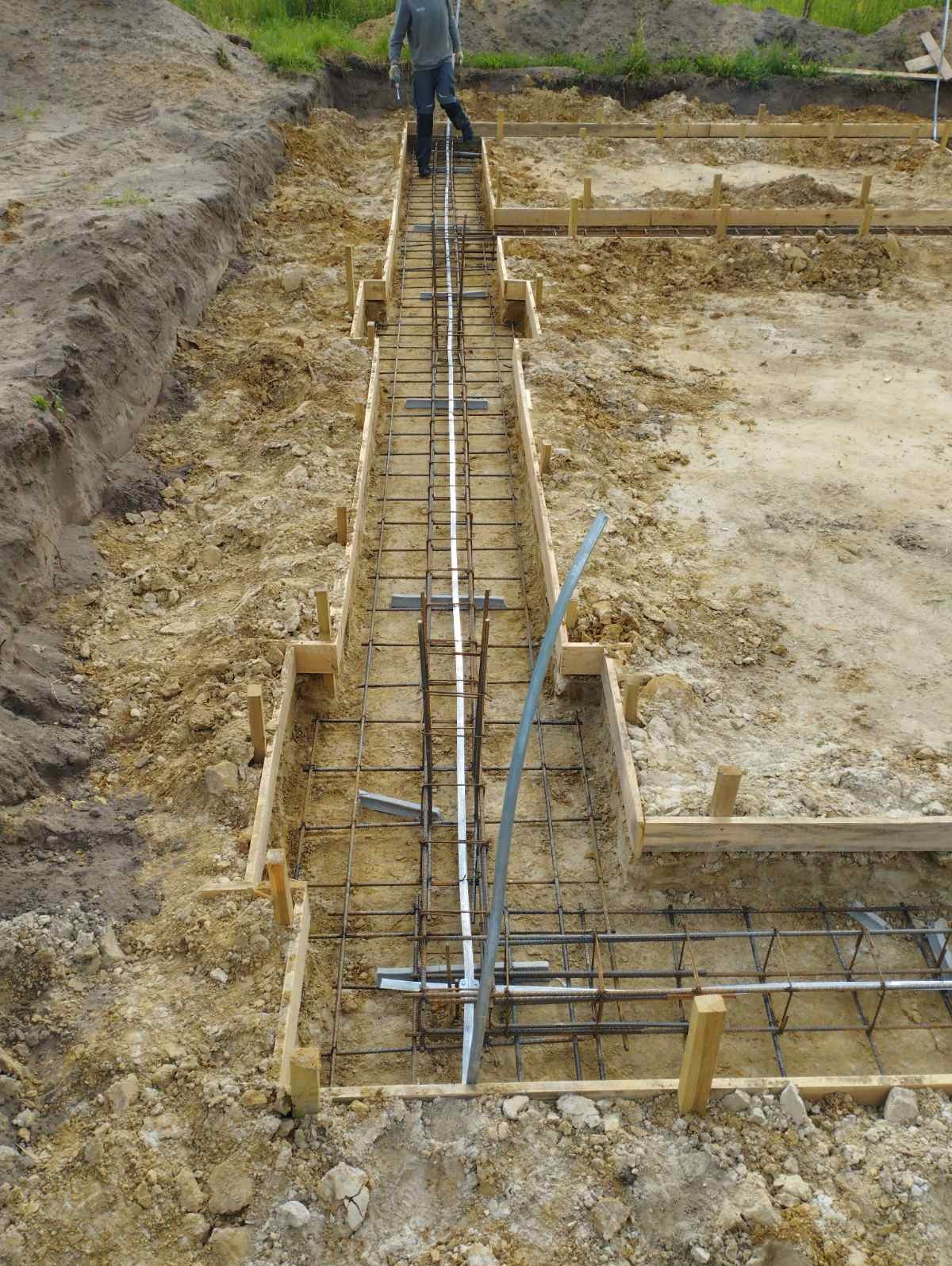Zbrojenie ławy fundamentowej w drewnianym szalunku, widok z góry na wykop z ziemią i pracownika w tle.