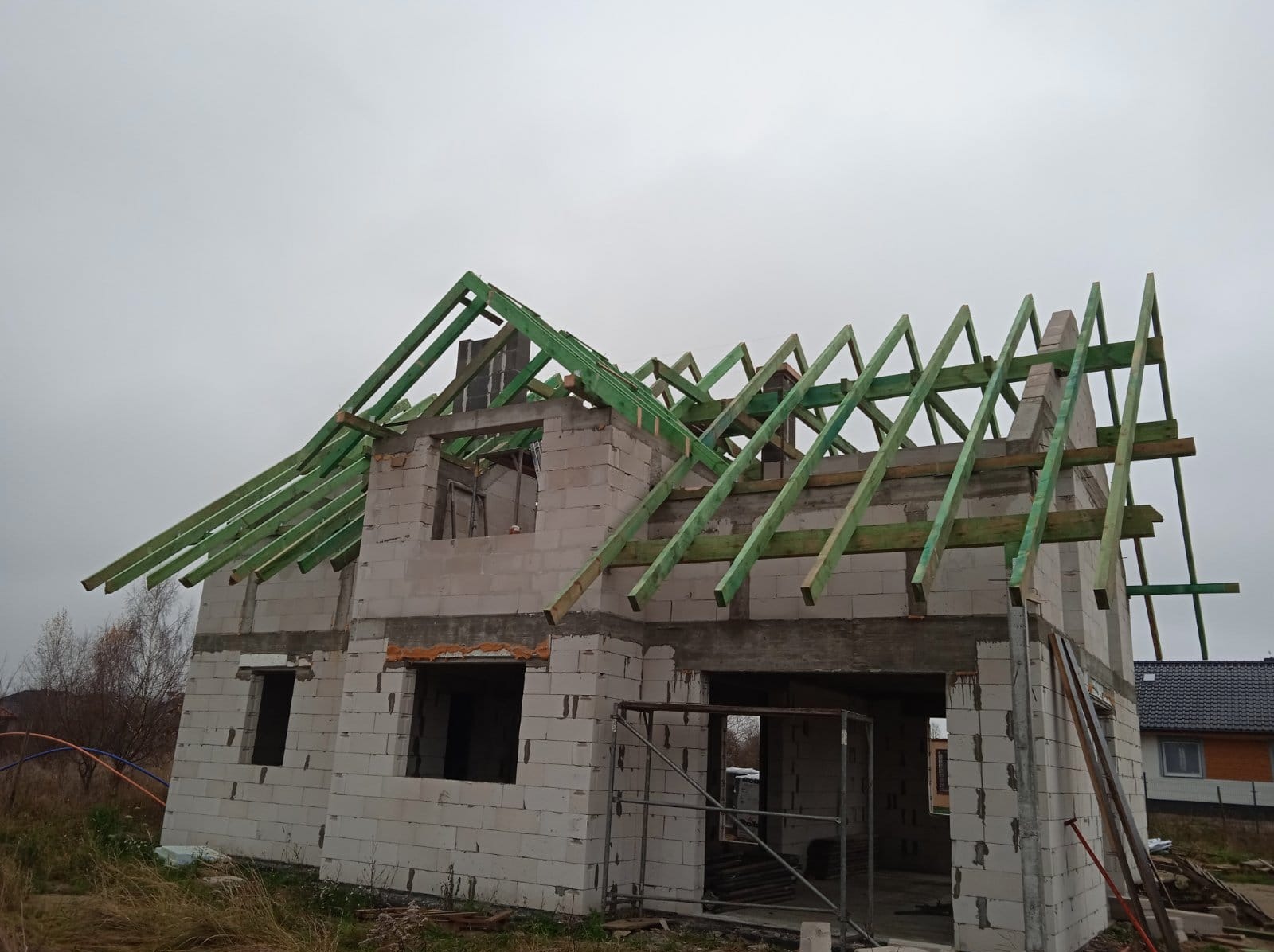 Surowy dom jednorodzinny w trakcie budowy, ściany z betonu komórkowego, widoczne zielone elementy konstrukcji dachu na tle pochmurnego nieba.