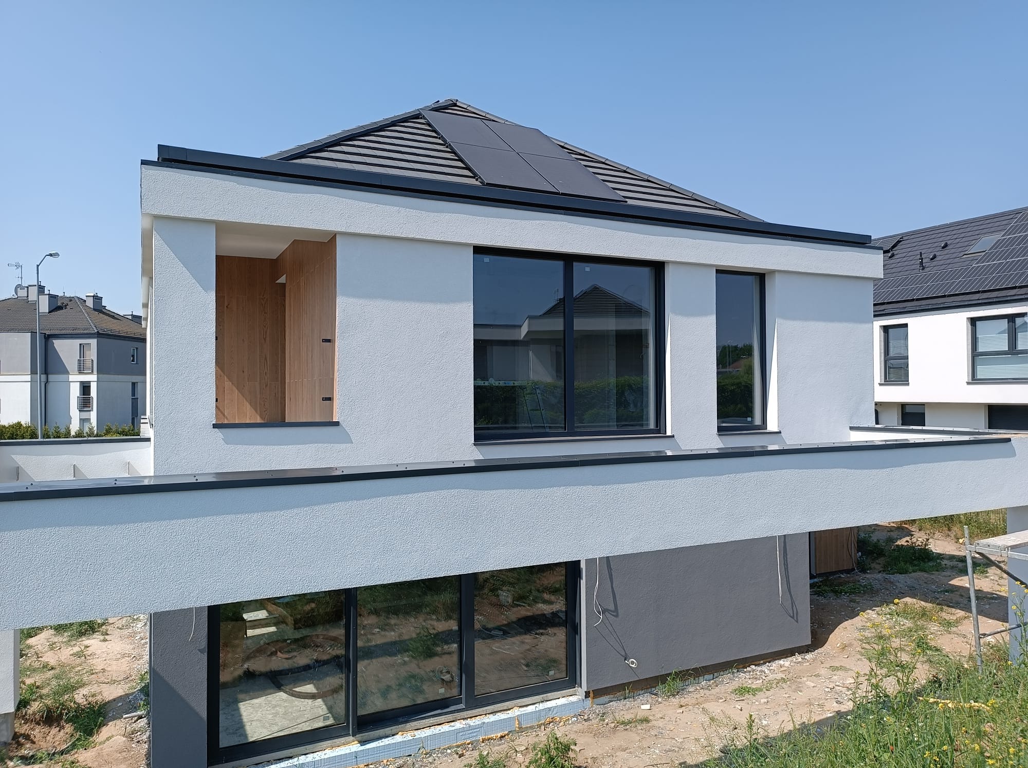 Nowoczesny dom z białą, chropowatą elewacją i panelami słonecznymi na dachu, widoczne okna z czarnymi ramami i surowe wnętrze bez wykończenia, w tle inne domy w podobnym stylu.