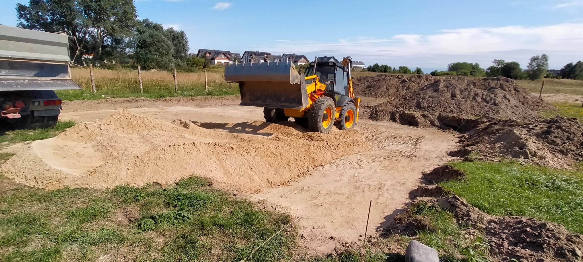 Żółta ładowarka na terenie budowy, wyrównująca piasek, z widocznymi śladami opon i stertami ziemi w tle, częściowo widoczna ciężarówka po lewej stronie, domy jednorodzinne w oddali.