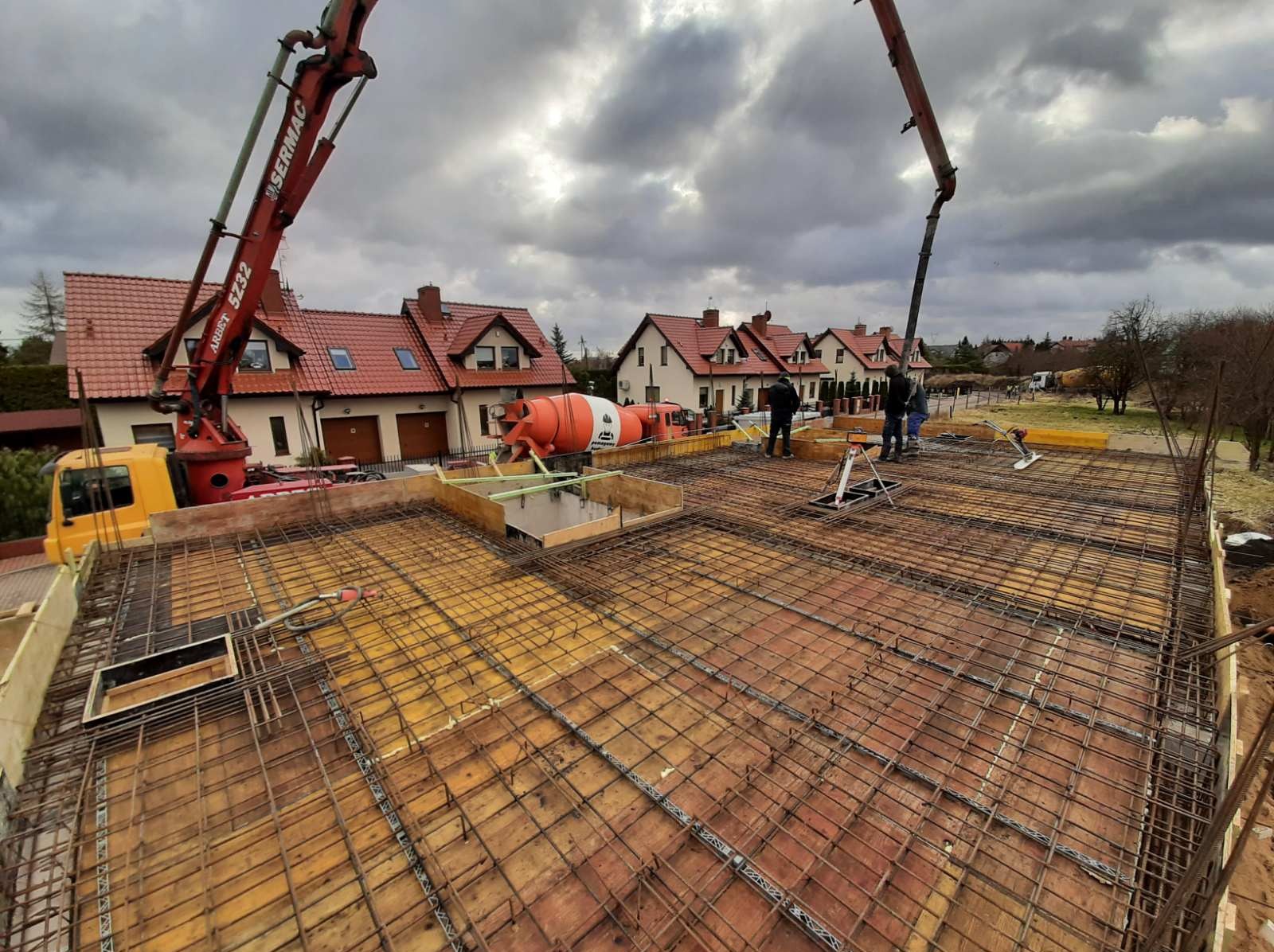 Zbrojenie płyty fundamentowej przygotowane do zalania betonem, widoczne pompy do betonu, mieszalnik i ekipa budowlana na tle domów jednorodzinnych.