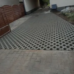 Szara, ażurowa kostka brukowa tworząca podjazd, płynnie przechodząca w tradycyjną kostkę. Widoczne elementy ogrodzenia i fragment budynku w tle.