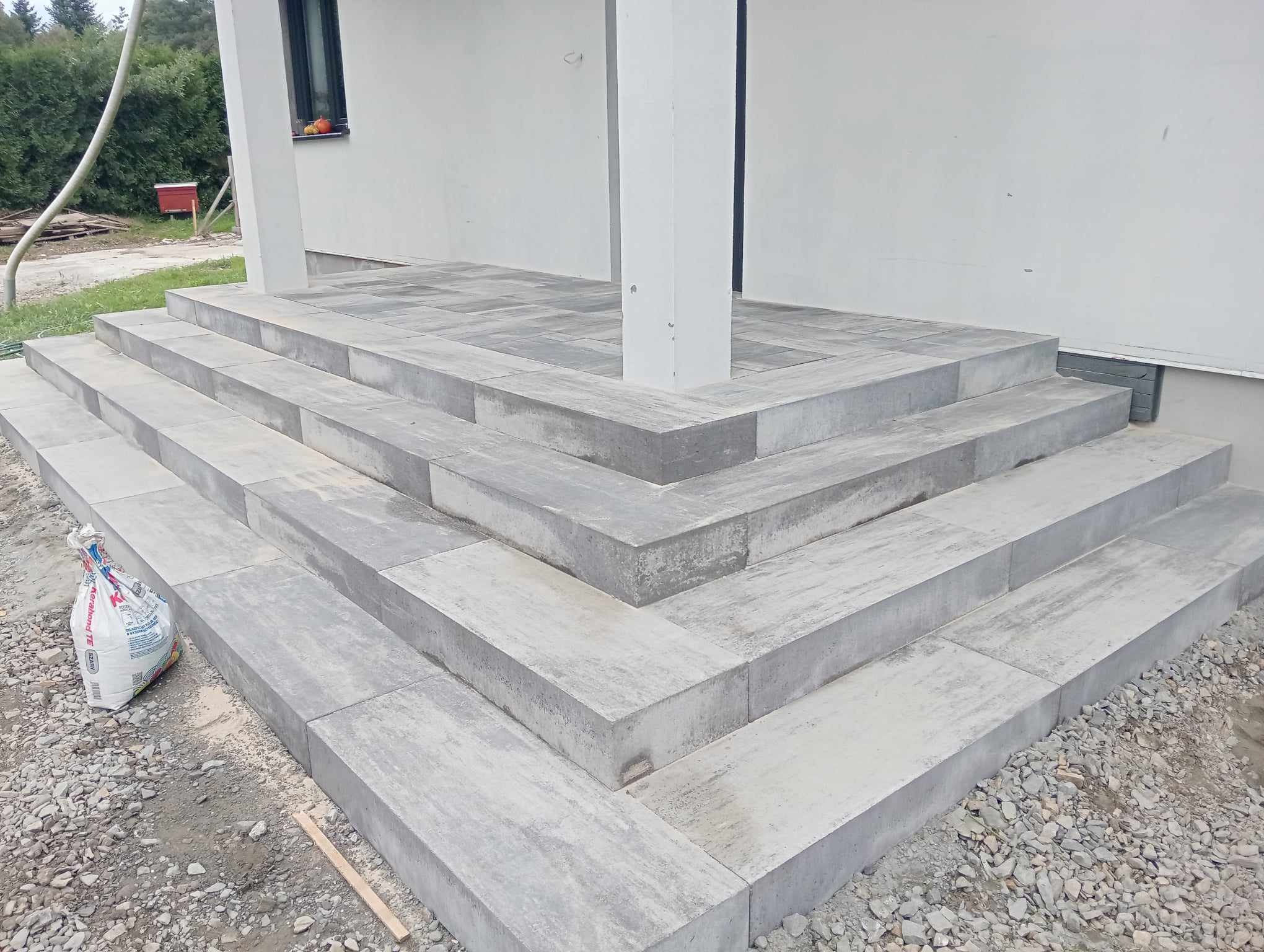 Schody wejściowe do domu wyłożone szarą kostką brukową, widoczny worek z zaprawą i drobne kamienie wokół, biały filar podtrzymujący zadaszenie.