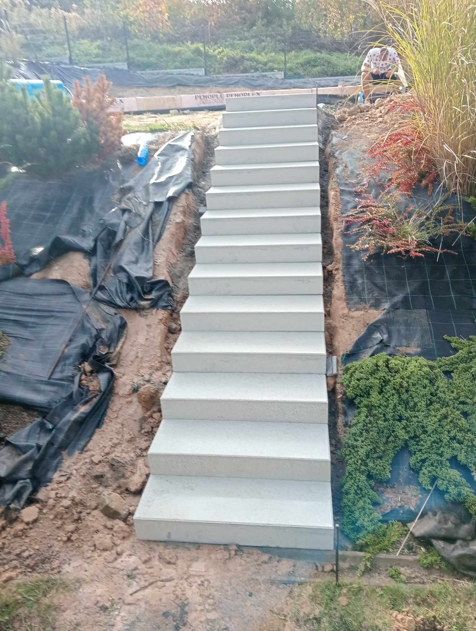 Betonowe schody ogrodowe prowadzące w górę zbocza, widoczne czarne maty szkółkarskie, fragment trawnika i roślinność ozdobna.