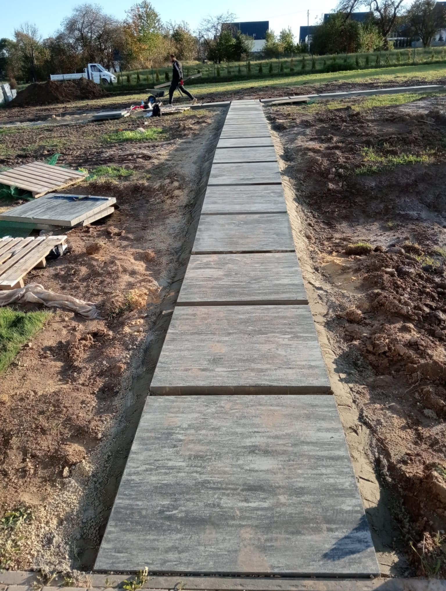 Ścieżka z płyt betonowych ułożona w ziemi, widoczny pracownik i samochód dostawczy w tle, teren przygotowany pod dalsze prace.