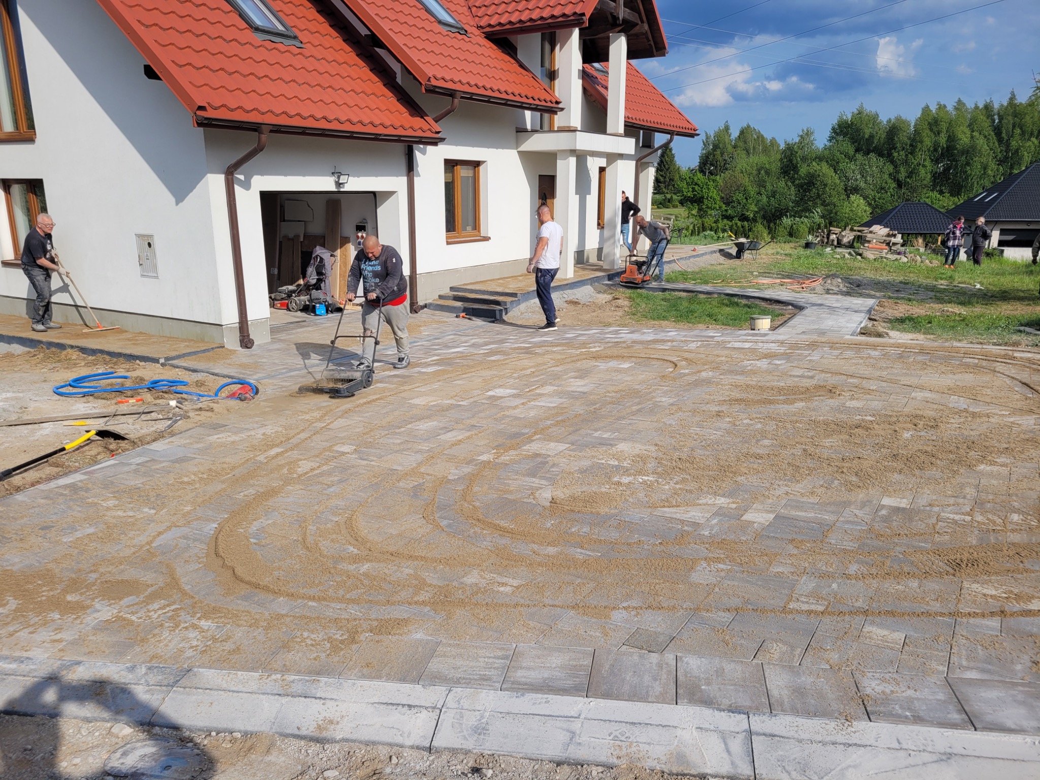 Układanie kostki brukowej na posesji domu jednorodzinnego. Widoczni pracownicy, narzędzia i częściowo ułożona kostka z piaskiem do wypełnienia szczelin.