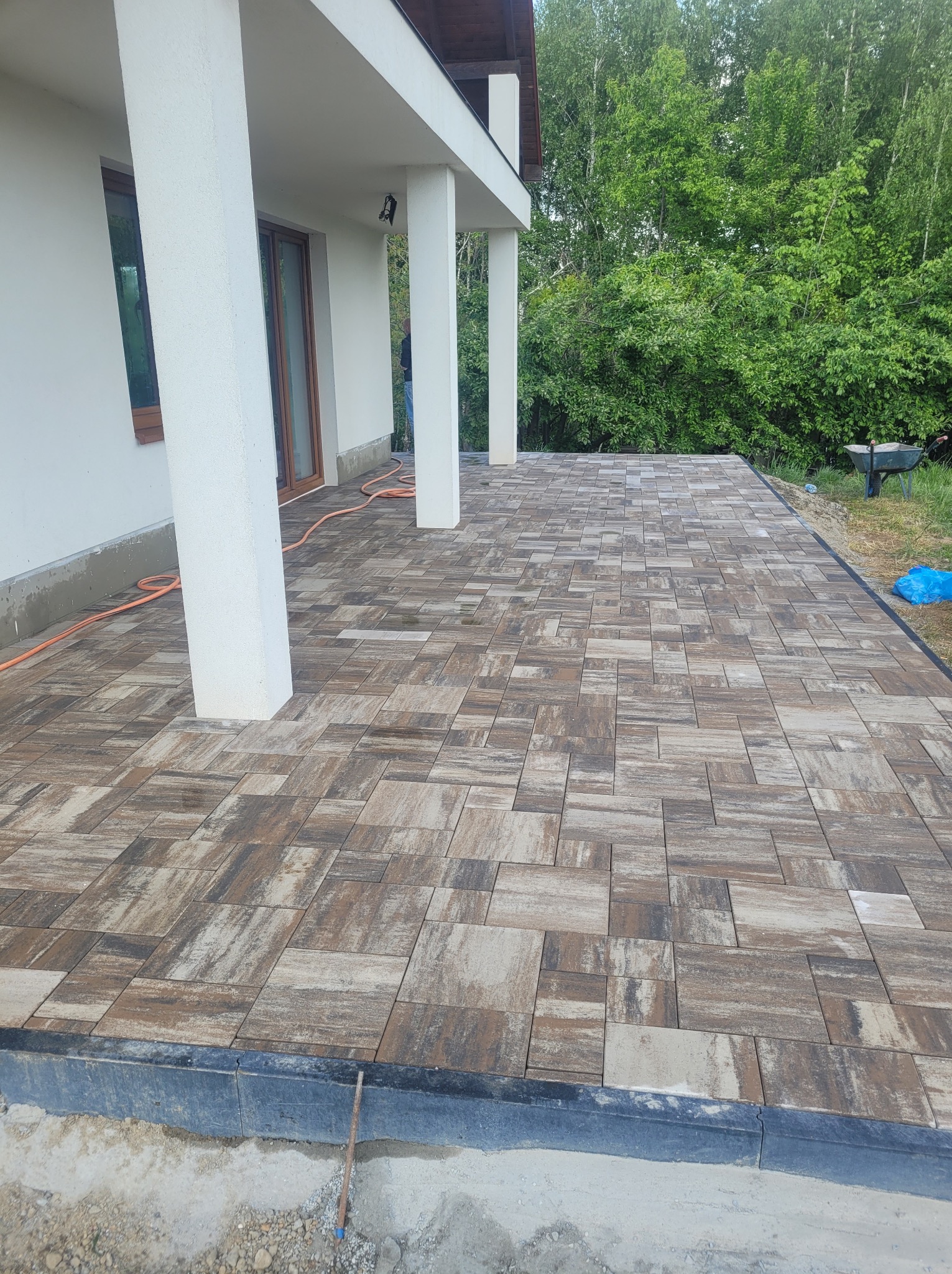 Ułożona kostka brukowa w odcieniach brązu i szarości na tarasie przy domu z białymi kolumnami. Widoczne zielone drzewa w tle.