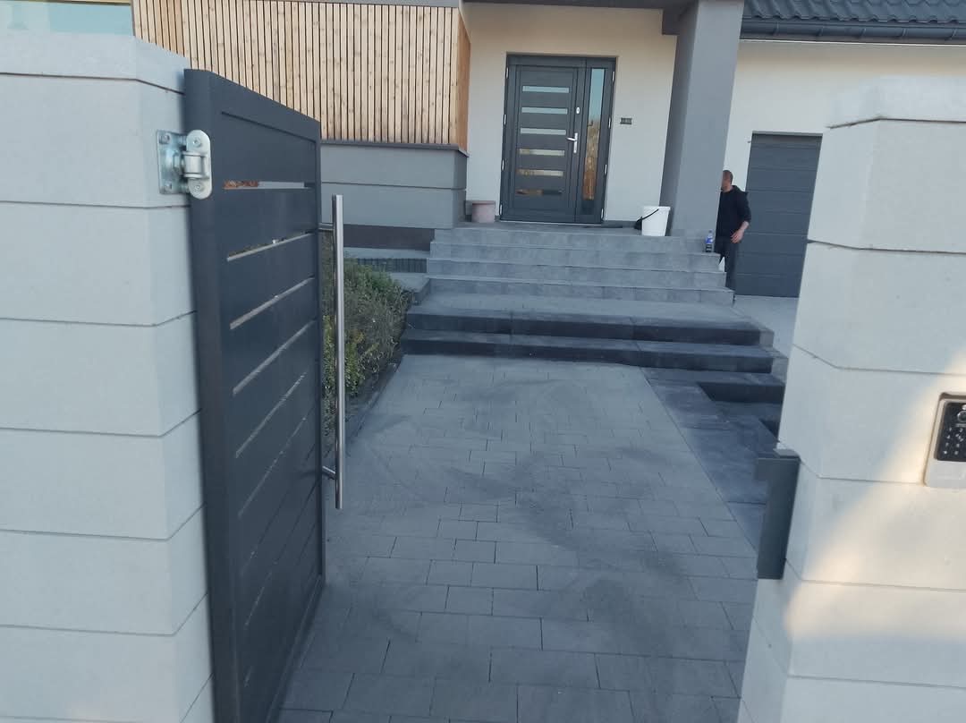 Nowoczesna posesja z szarą kostką brukową, metalową furtką i jasnymi słupkami. Schody wejściowe również wyłożone kostką. Minimalistyczny design.