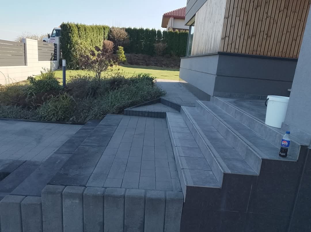 Nowoczesne schody i chodnik z szarej kostki brukowej prowadzące do domu z drewnianą elewacją. Widoczny fragment ogrodzenia i zadbany trawnik.