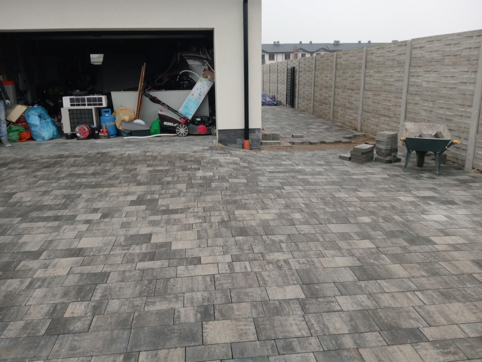 Ułożona kostka brukowa w Rzeszowie, widoczny fragment ogrodzenia i garaż z otwartą bramą. Kostka w odcieniach szarości. Częściowo ułożona nawierzchnia.