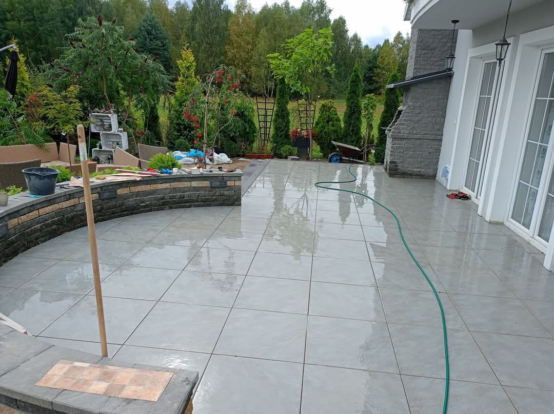 Ułożona, szara, błyszcząca nawierzchnia z płytek na tarasie domu, z elementami wykończeniowymi z kamienia. W tle zielony ogród i fragment elewacji budynku.