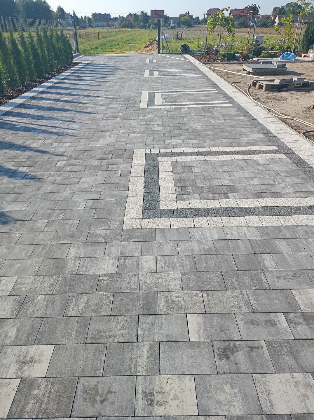 Nowoczesna, szara kostka brukowa z dekoracyjnymi wstawkami w kształcie prostokątów, otoczona zielenią i ogrodzeniem. Widok z perspektywy.