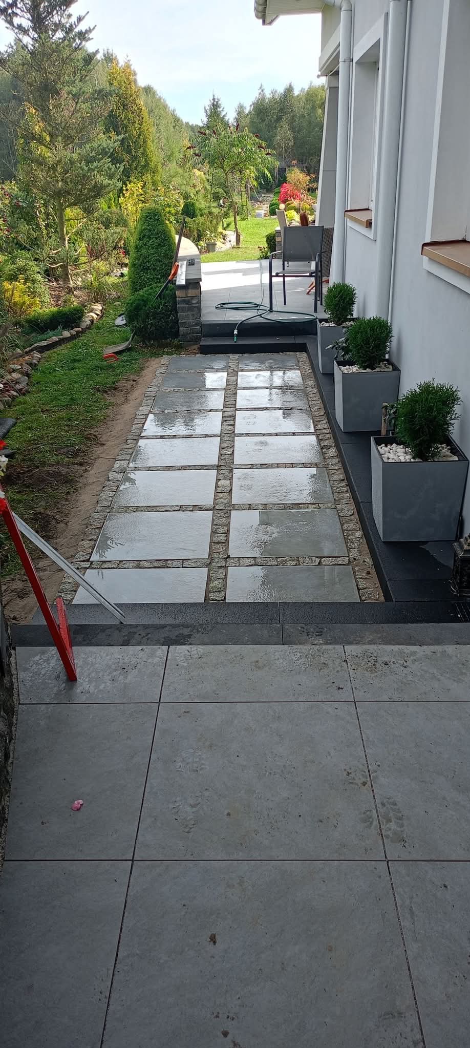 Ułożona ścieżka z kwadratowych płyt granitowych oddzielonych kostką, prowadząca od schodów do drzwi domu, obok donice z zielenią.