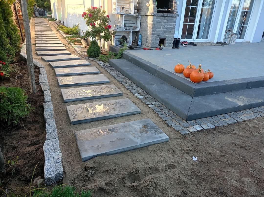 Ścieżka z płyt betonowych prowadząca do domu, obrzeża z kostki granitowej, jesienna dekoracja z dyń. Widok na ogród i elewację domu.