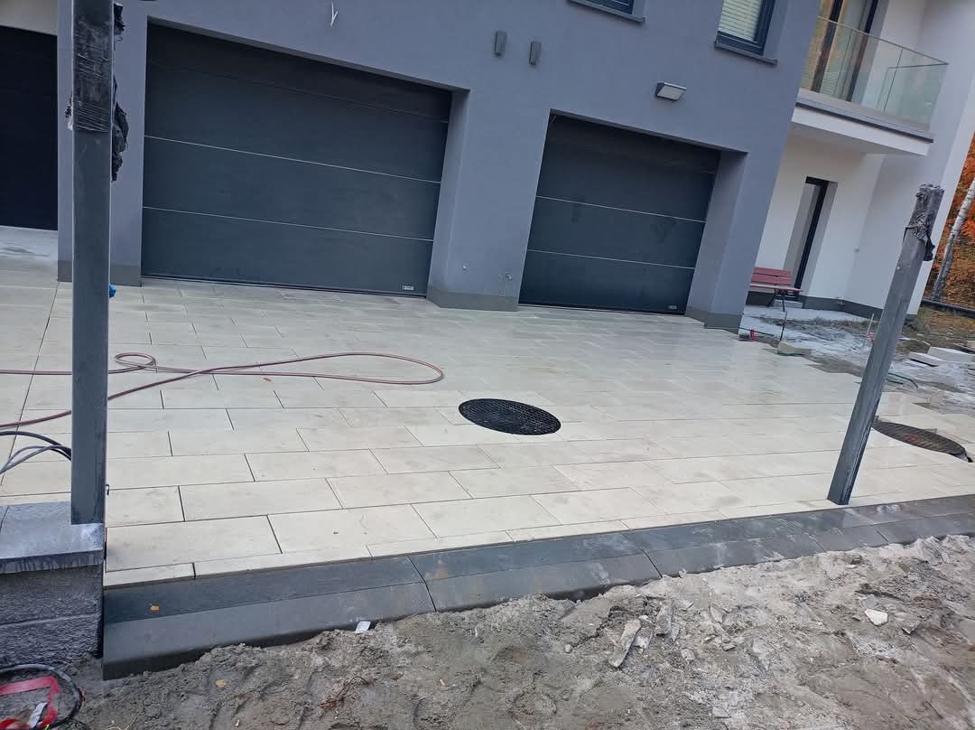 Jasna kostka brukowa ułożona przed nowoczesnym domem z szarymi bramami garażowymi. Widoczne obrzeża i niedokończone prace ziemne, z elementami instalacji ogrodzeniowej.