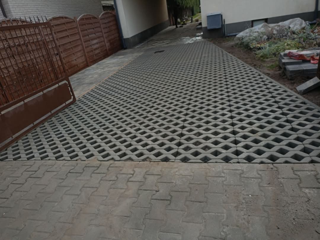 Szara, ażurowa kostka brukowa tworząca podjazd, płynnie przechodząca w tradycyjną kostkę. Widoczne elementy ogrodzenia i fragment budynku w tle.