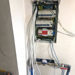 W KONTAKCIE ROBERT ANZORGE - Rozdzielnica elektryczna z licznymi bezpiecznikami i przewodami w surowym wnętrzu, widoczne także rury instalacji wodnej i narzędzia.