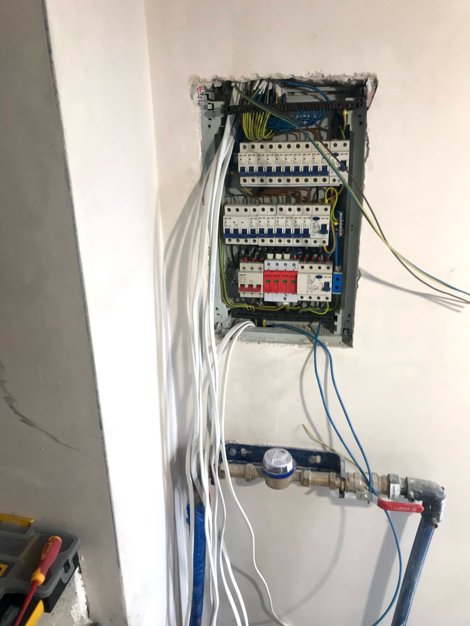 Rozdzielnica elektryczna z licznymi bezpiecznikami i przewodami w surowym wnętrzu, widoczne także rury instalacji wodnej i narzędzia.