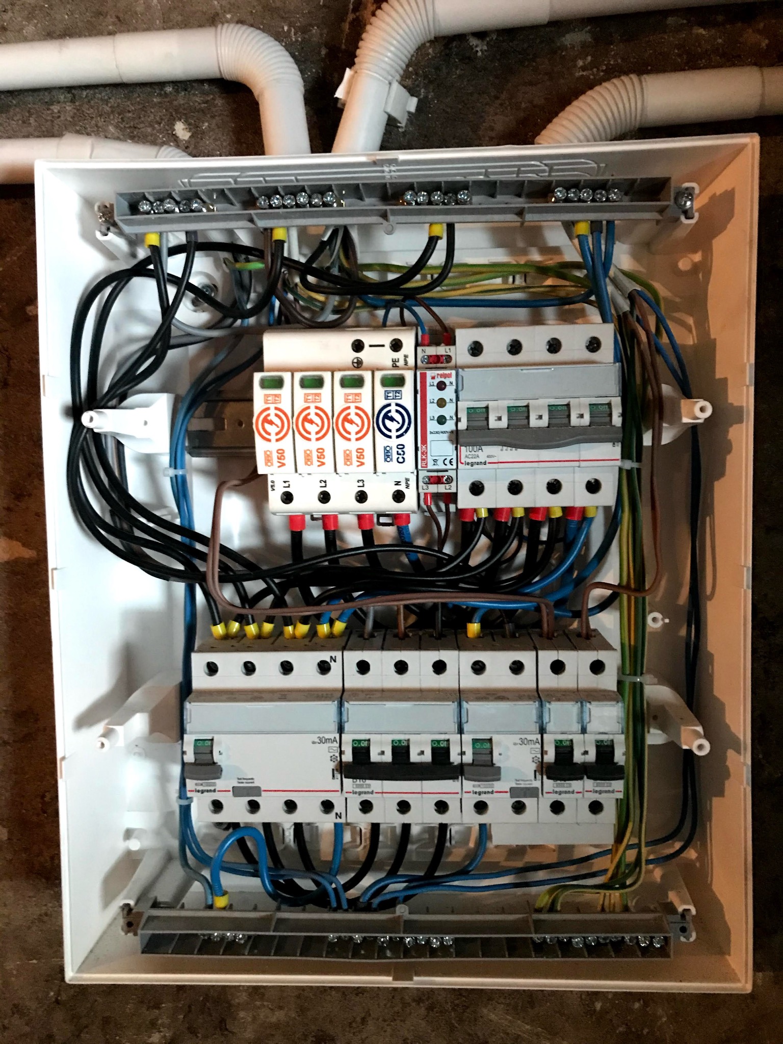 Rozdzielnica elektryczna w białej obudowie z widocznymi bezpiecznikami, przewodami i elementami montażowymi. Instalacja elektryczna w trakcie prac.