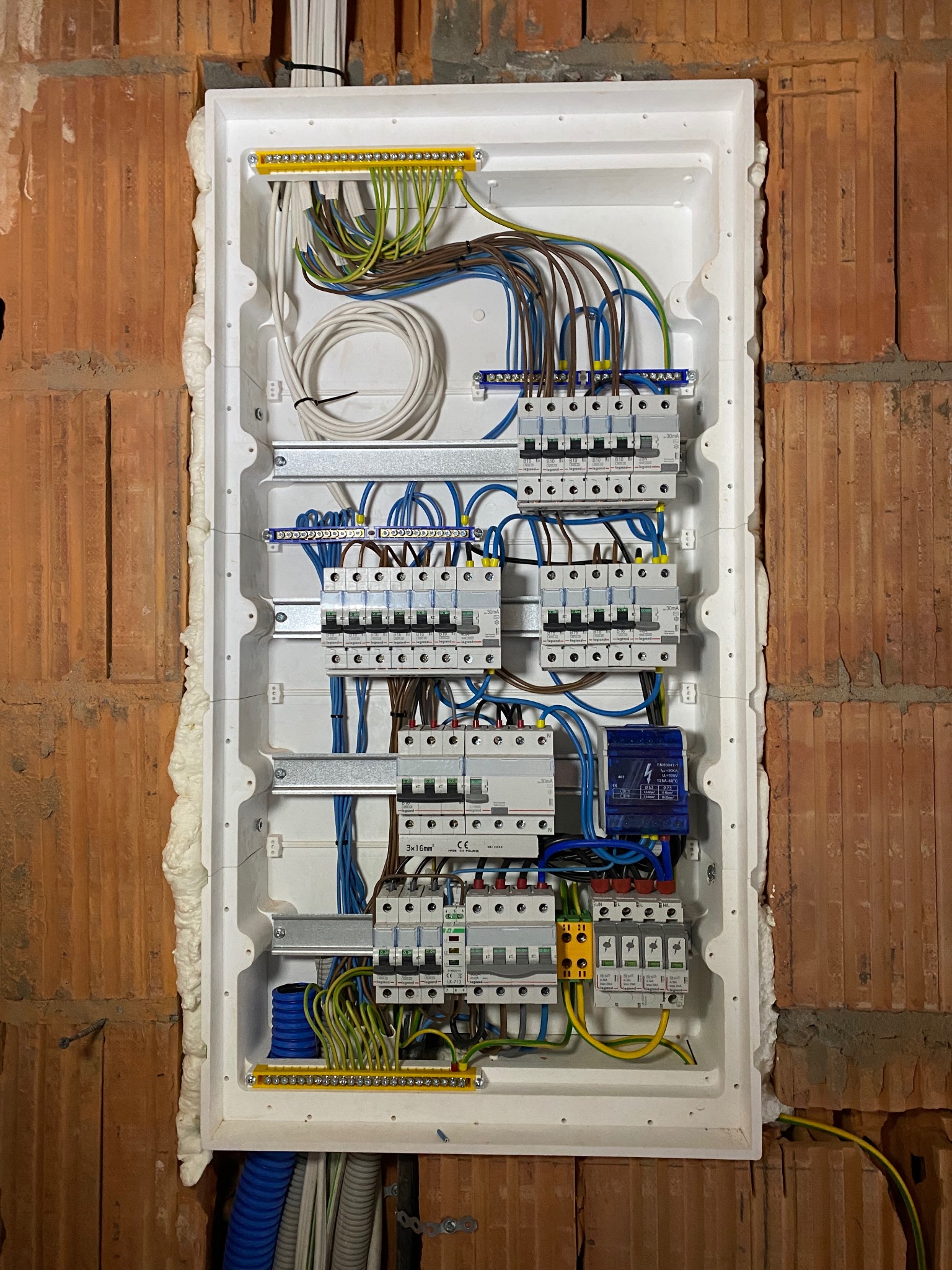 Rozdzielnica elektryczna wmurowana w ścianę z cegły, zabezpieczona pianką montażową. Widoczne kable, bezpieczniki i inne elementy instalacji elektrycznej.